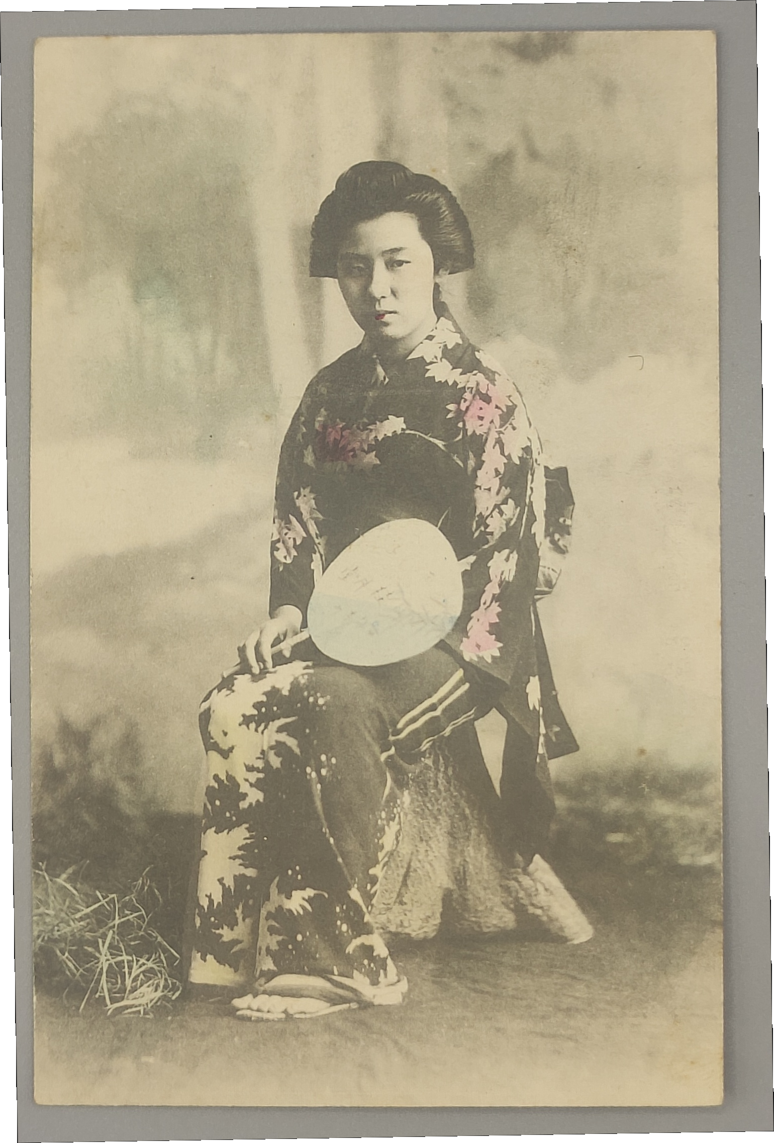 Front of a Meiji-era hand-tinted postcard: woman in kimono holding an uchiwa fan, outdoors